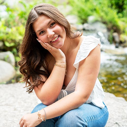 Smiling young woman sits outdoors on a rock, hand on cheek, with green foliage and a stream.