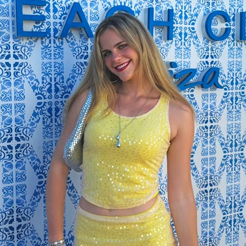 Smiling student in yellow sequined outfit, silver bag, against blue and white patterned wall.